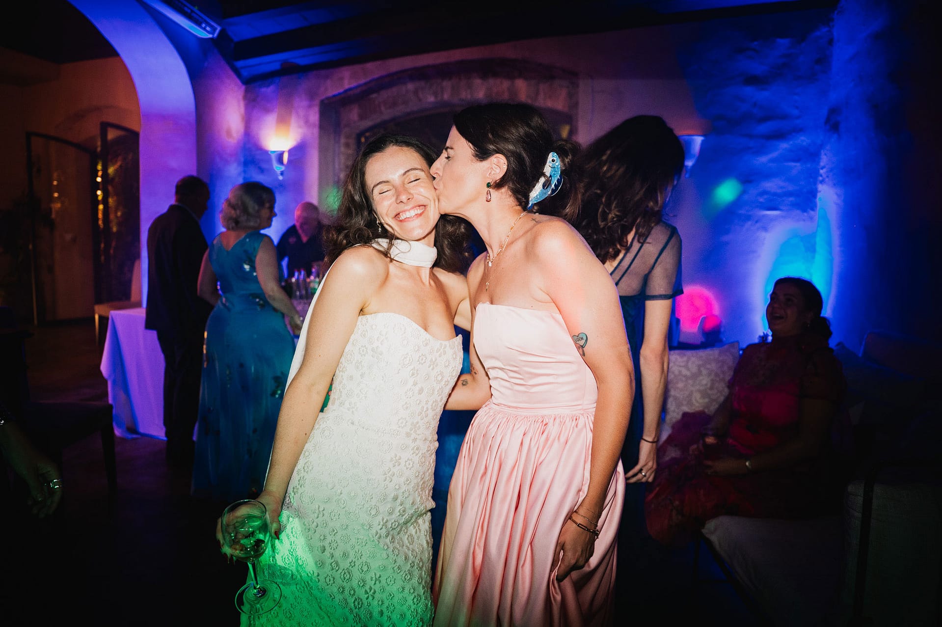 Two women embrace and kiss at a lively wedding reception with colorful lighting. - Barcelona Wedding Photographer