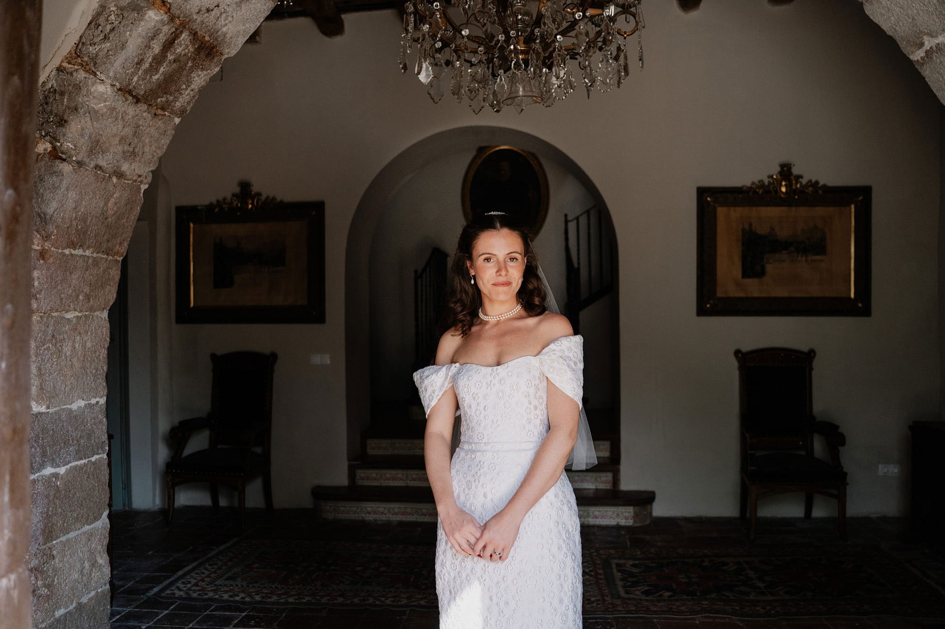 Bride in a white dress stands in an arched entryway with a chandelier overhead. - Barcelona Wedding Photographer
