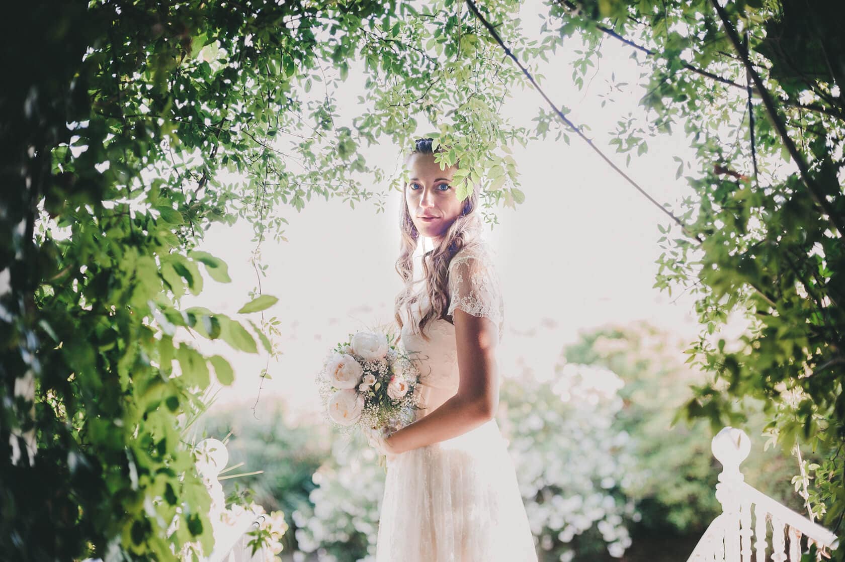 Bride in a lace dress holding a bouquet, framed by lush greenery in Barcelona - Barcelona Wedding Photographer