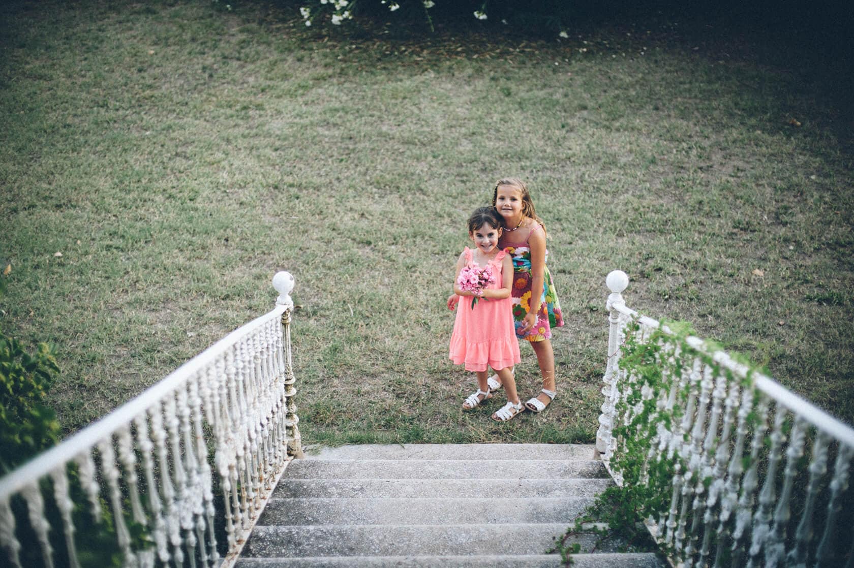 Two young girls in dresses stand on stairs, one holding a bouquet, in a garden setting. - Barcelona Wedding Photographer