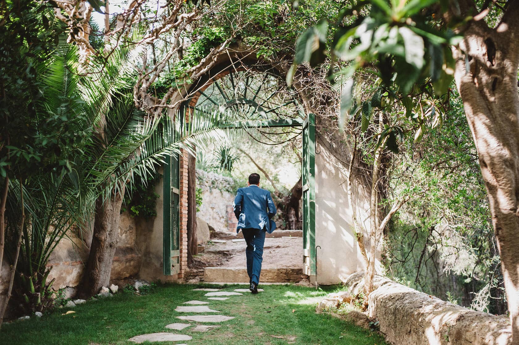 Groom in blue suit walks through garden archway at Villa Almanita, Sitges destination wedding. - Barcelona Wedding Photographer