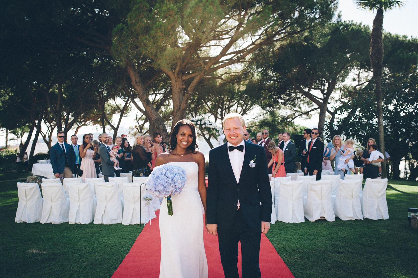 Newlyweds Tsega and Anders smiling on a red carpet in S'Agaro, Spain. - Barcelona Wedding Photographer