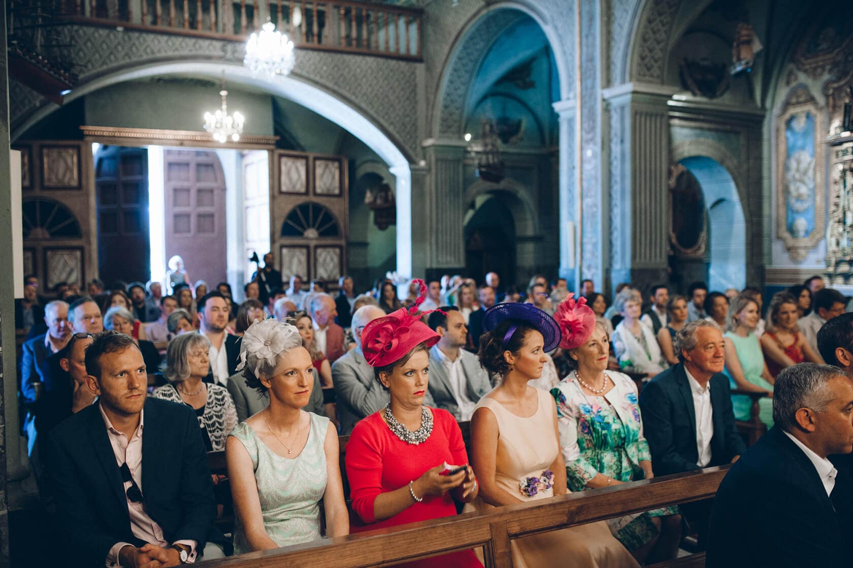 Wedding guests in ornate hats seated in a church during a Casanova Estate wedding. - Barcelona Wedding Photographer