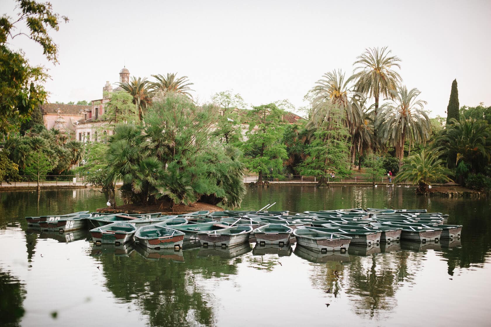 Boats docked in a serene lake with lush greenery and a historic building in Barcelona. - Barcelona Wedding Photographer