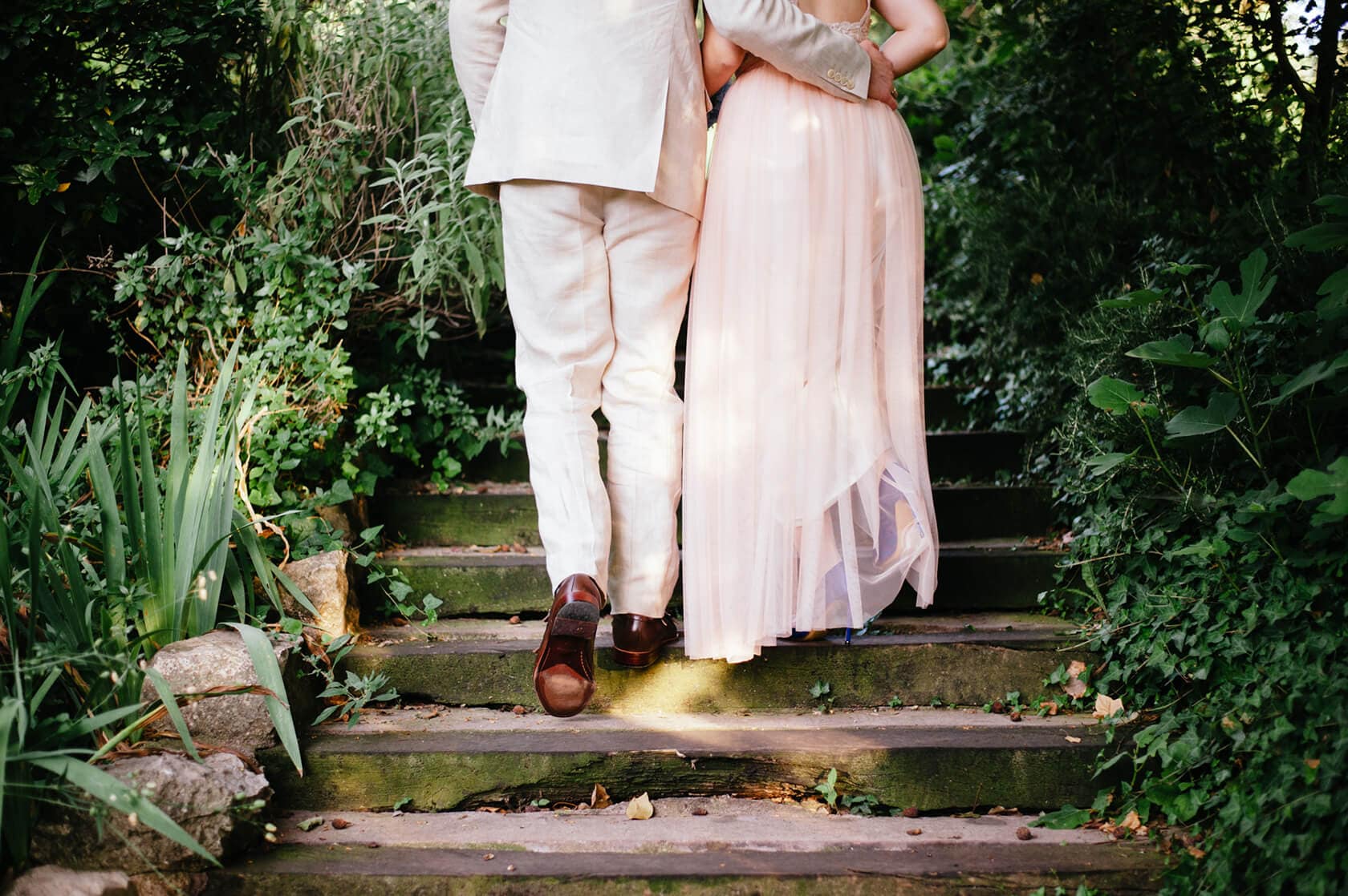 Couple in vintage style wedding attire walking up stone steps surrounded by greenery - Barcelona Wedding Photographer