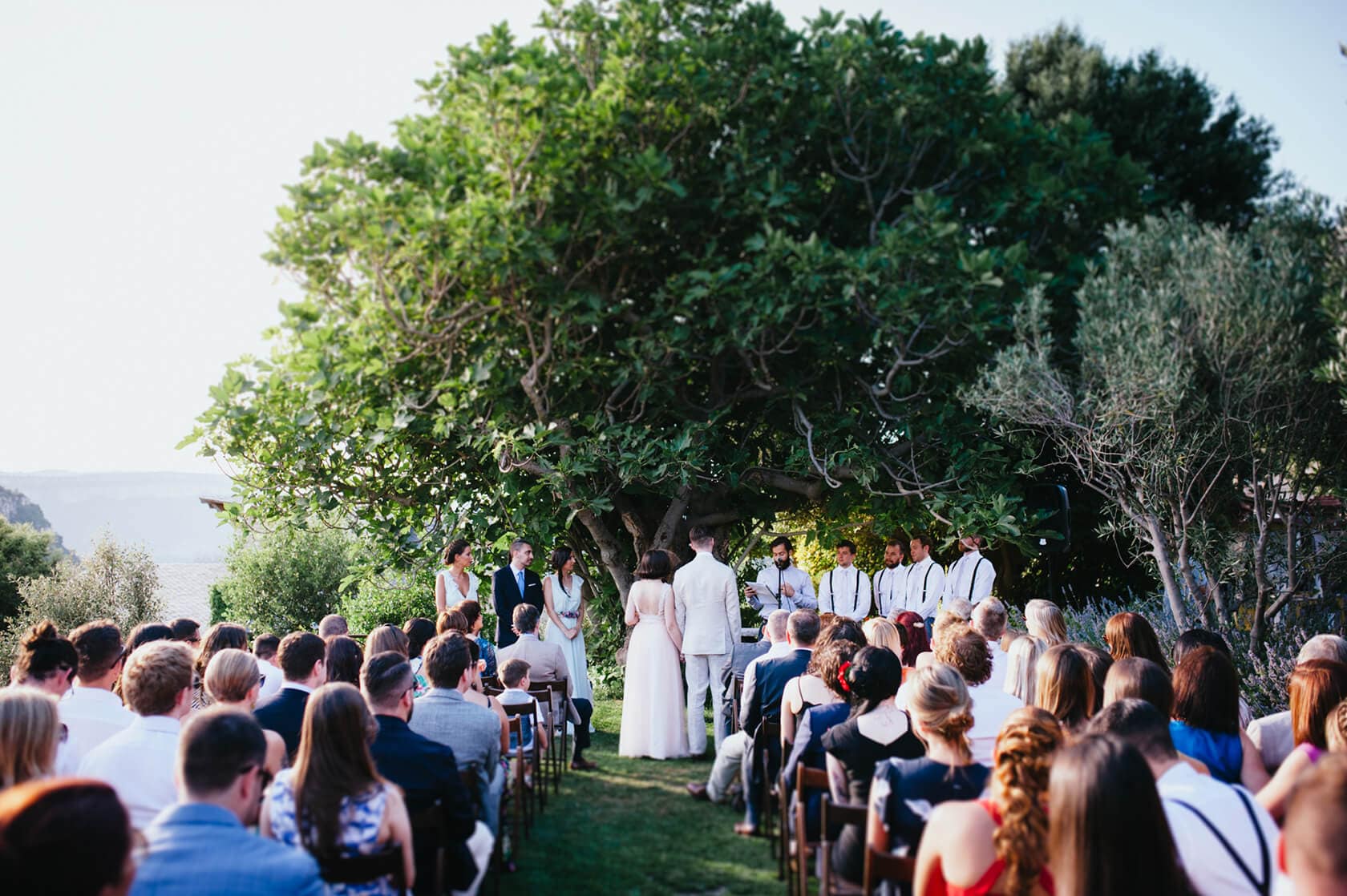 A vintage style wedding ceremony outdoors under a large tree with guests seated. - Barcelona Wedding Photographer