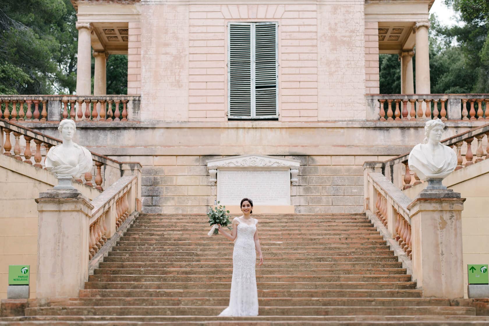 Bride in a white lace wedding dress holding a bouquet on the steps of Parc del Laberint d'Horta. - Barcelona Wedding Photographer
