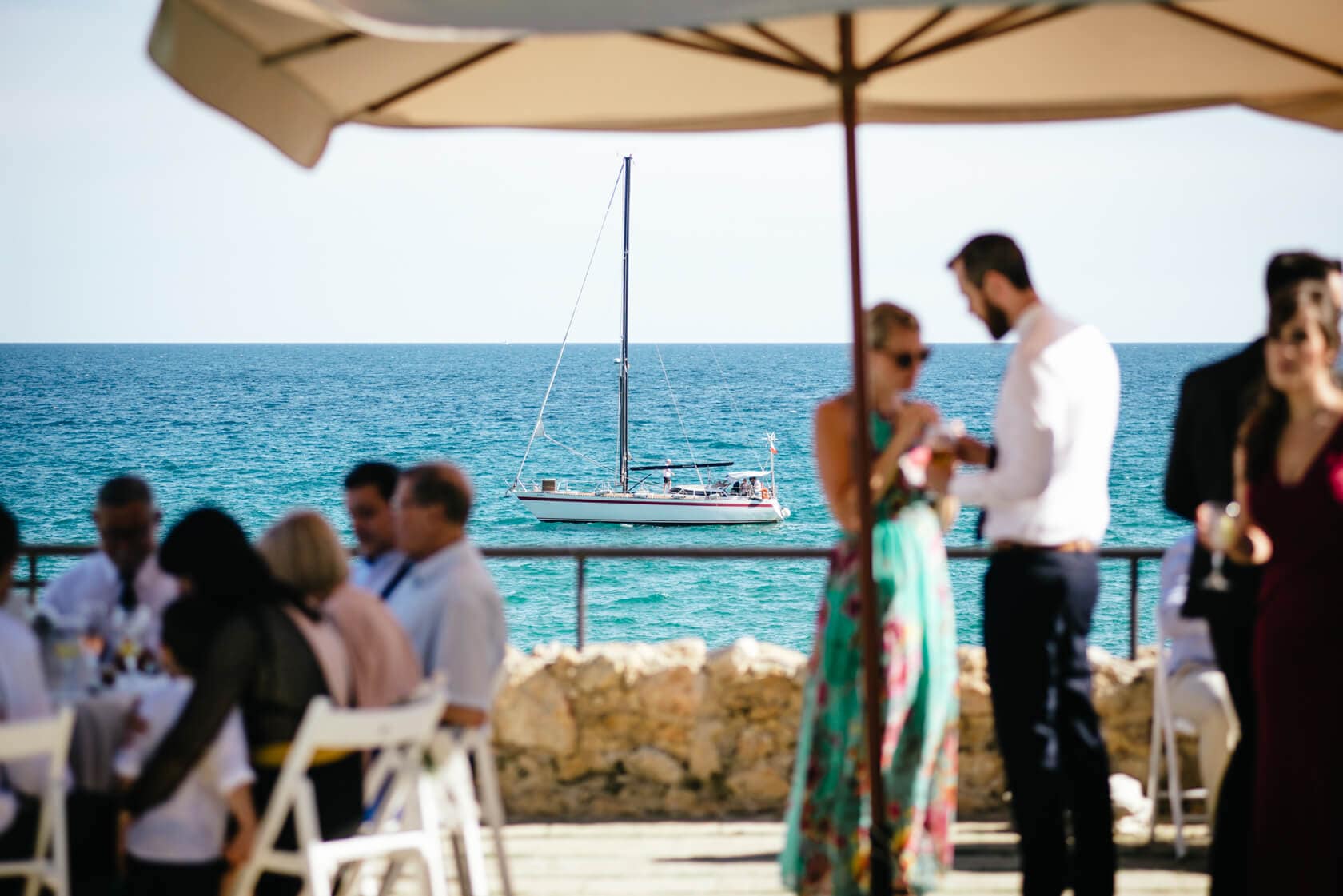 Wedding guests at Castell de Tamarit, Sitges, with a sailboat in the sea - Barcelona Wedding Photographer
