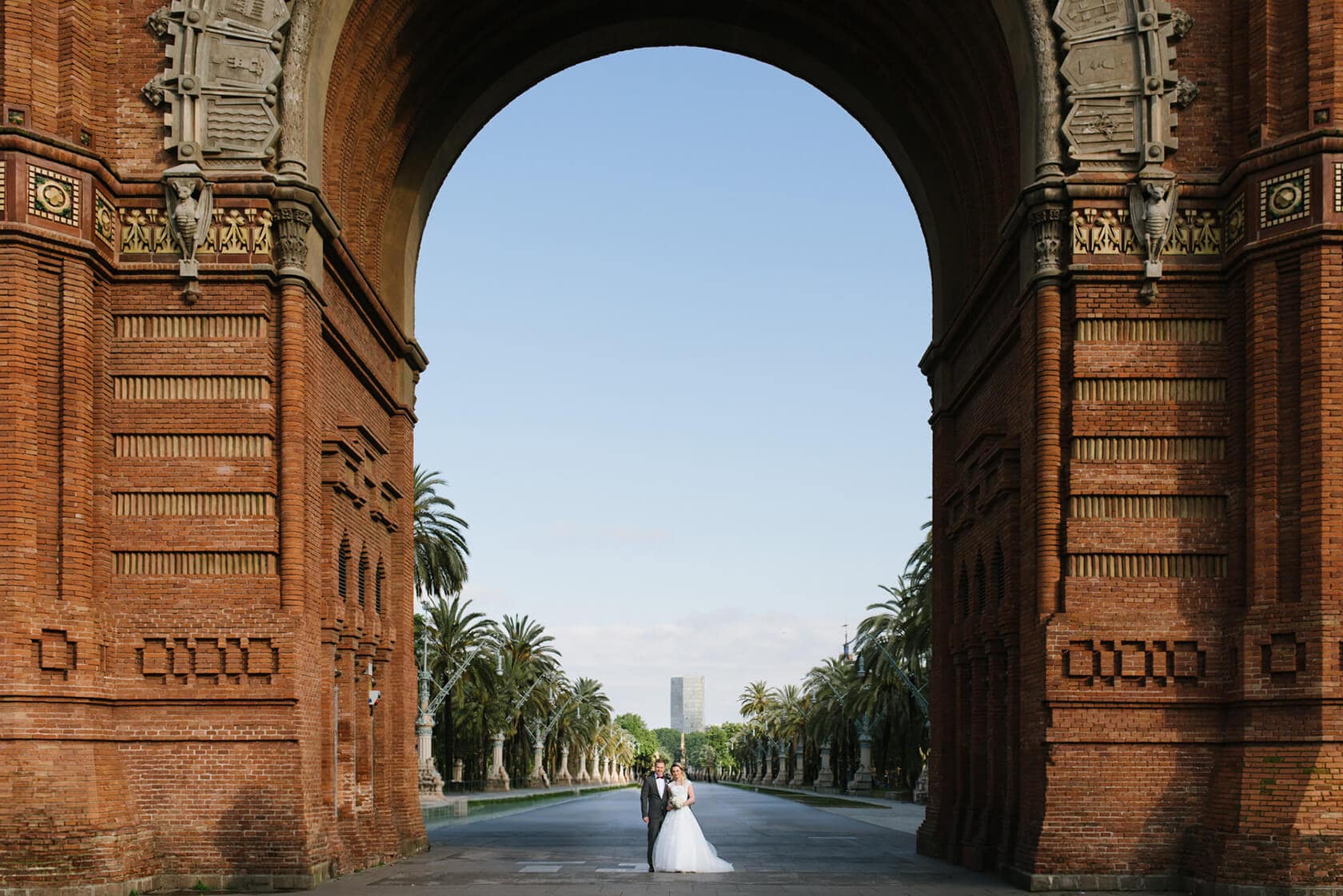 Bride and groom in wedding attire framed by Arc de Triomf in Barcelona city. Barcelona destination wedding.