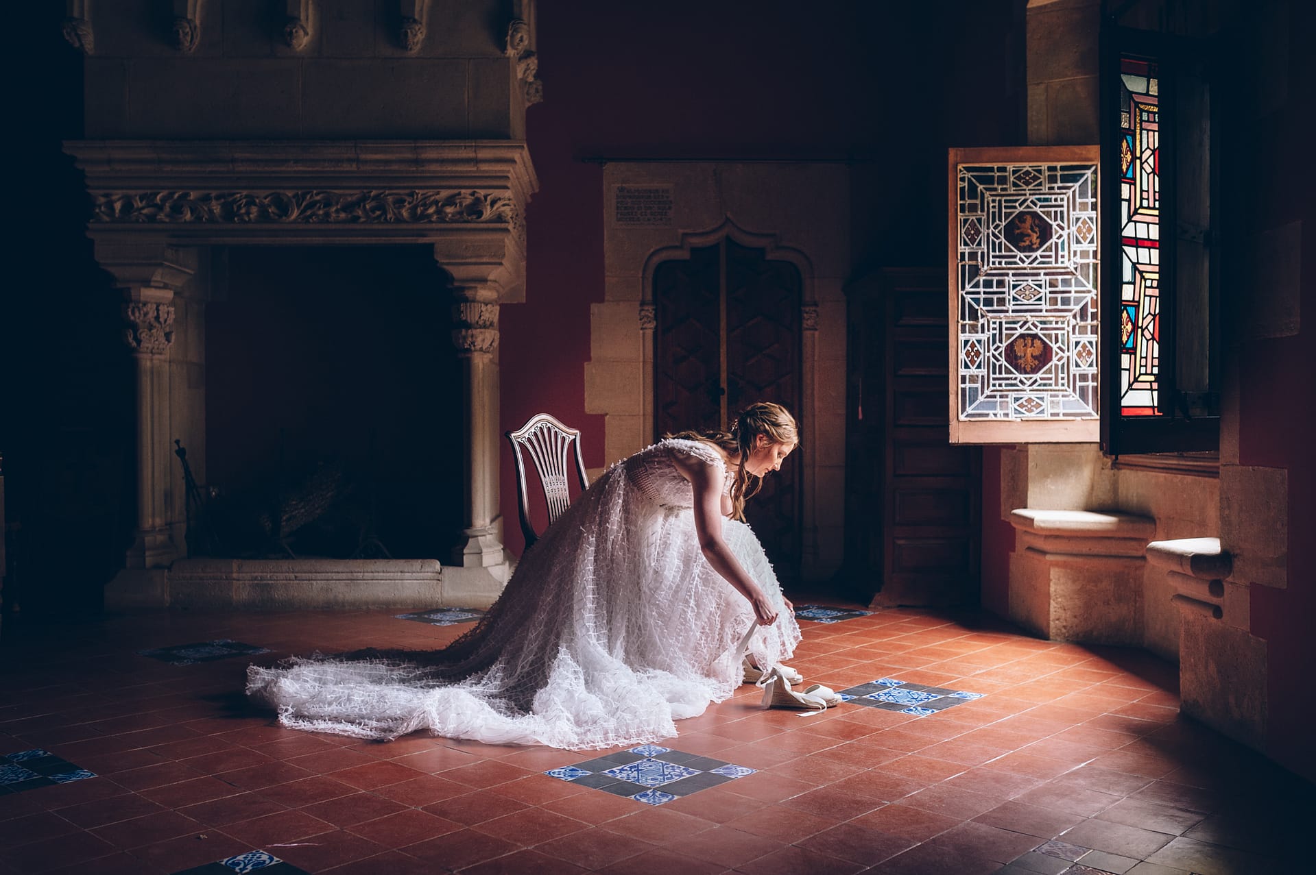 Bride in a white gown bends down to put on her shoes in a grand room with stained glass. - Barcelona Wedding Venues